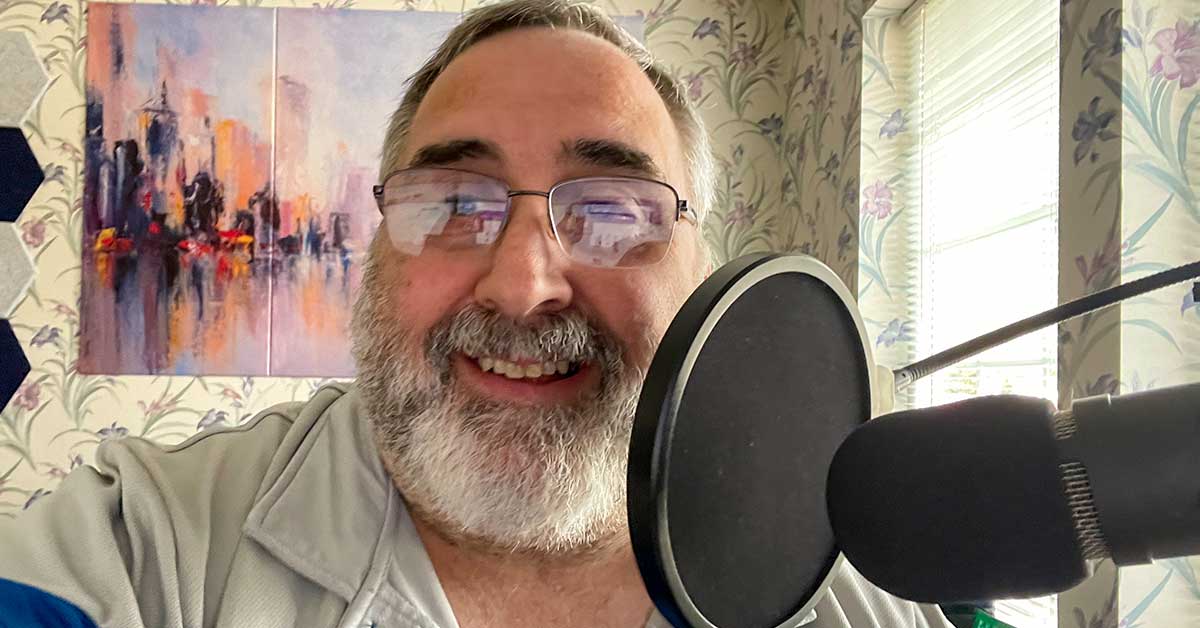 Ed Bejarana smiling behind a professional microphone setup, with a colorful abstract painting and floral wallpaper in the background, capturing a warm and approachable podcasting environment.