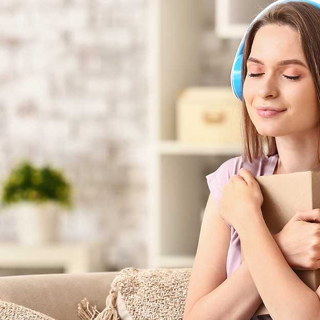 Young woman sitting on a cozy sofa, wearing blue headphones and hugging a book with a peaceful smile, symbolizing the joy of listening to audiobooks in a serene home environment.